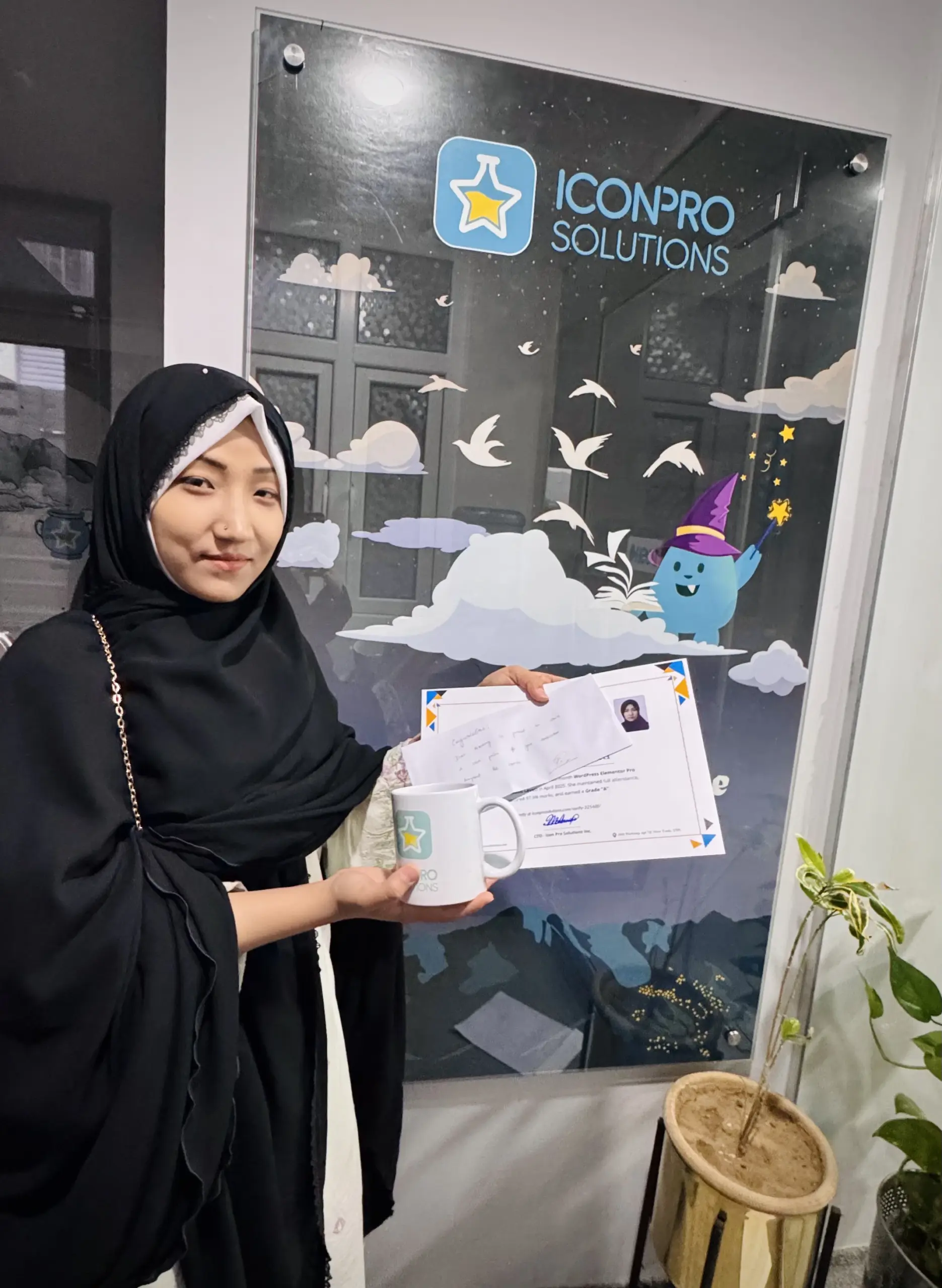 A woman in a black hijab and a black dress holds a white mug and a certificate in front of a glass door with a website