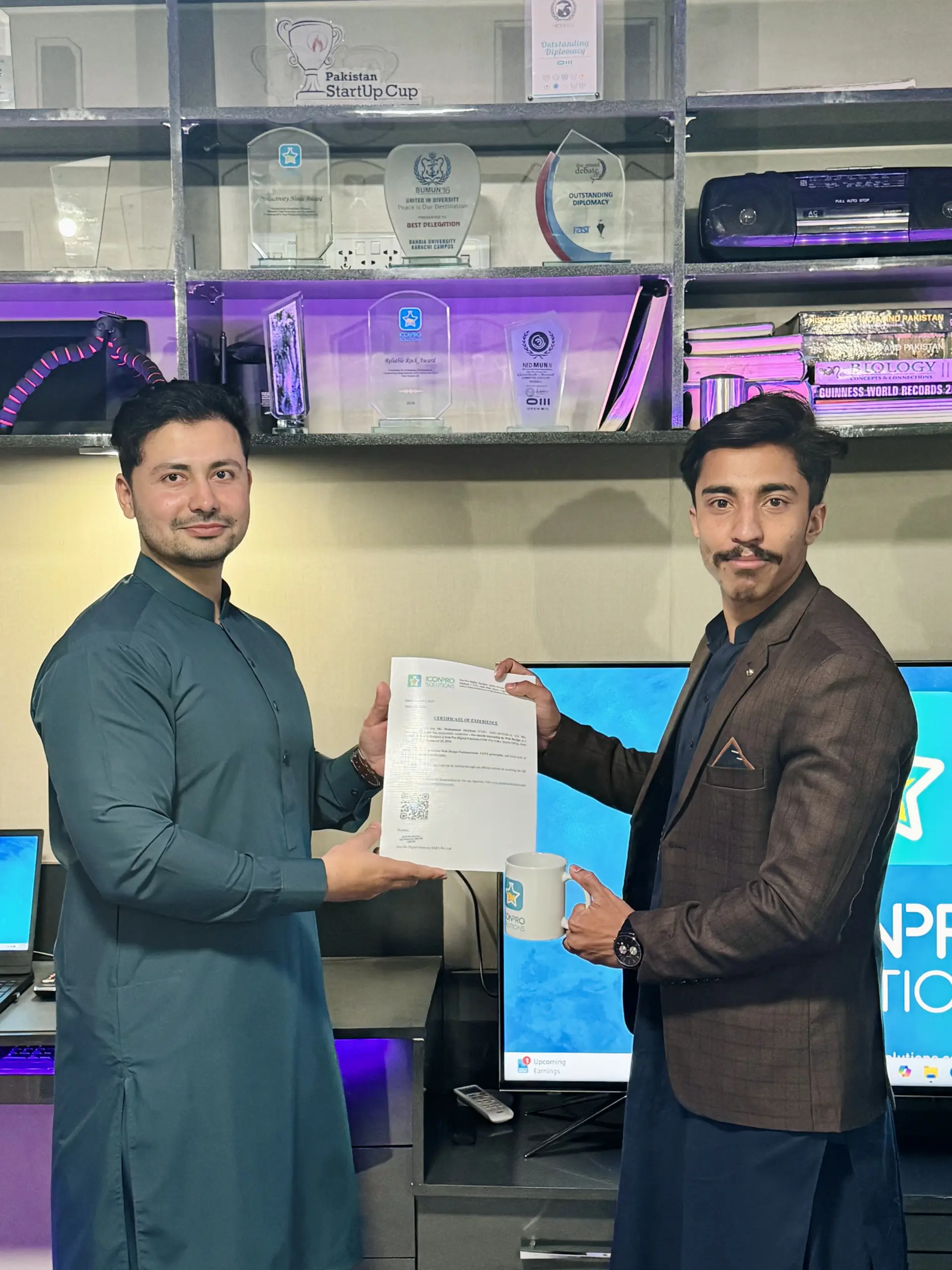 Two men in traditional Pakistani clothing holding a document in front of a wall of awards and a TV screen with a website