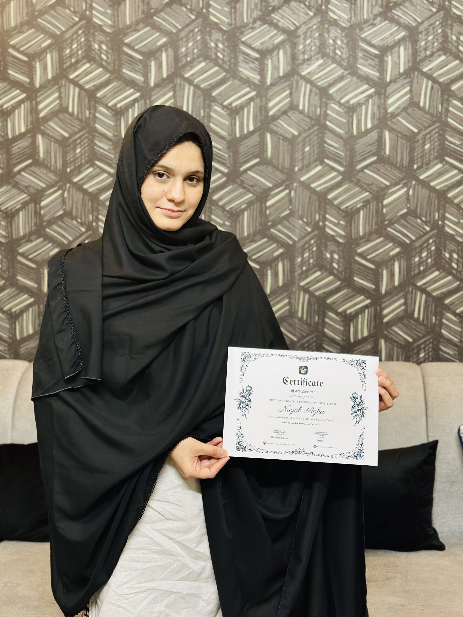website design A woman in a black hijab and white clothing holds a certificate in front of a patterned wall
