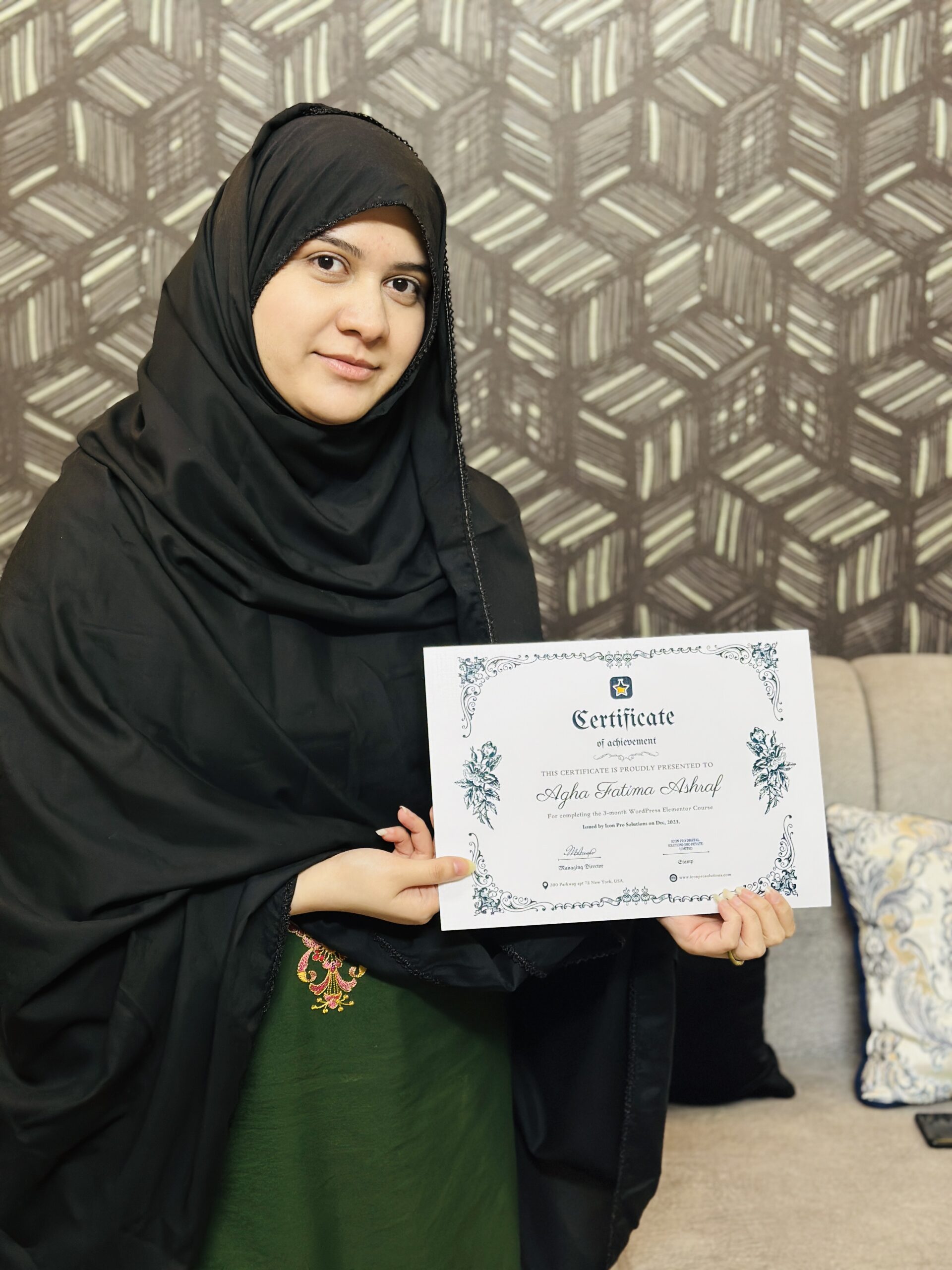 website design A woman in a black hijab and green dress holding a certificate in front of a patterned wall