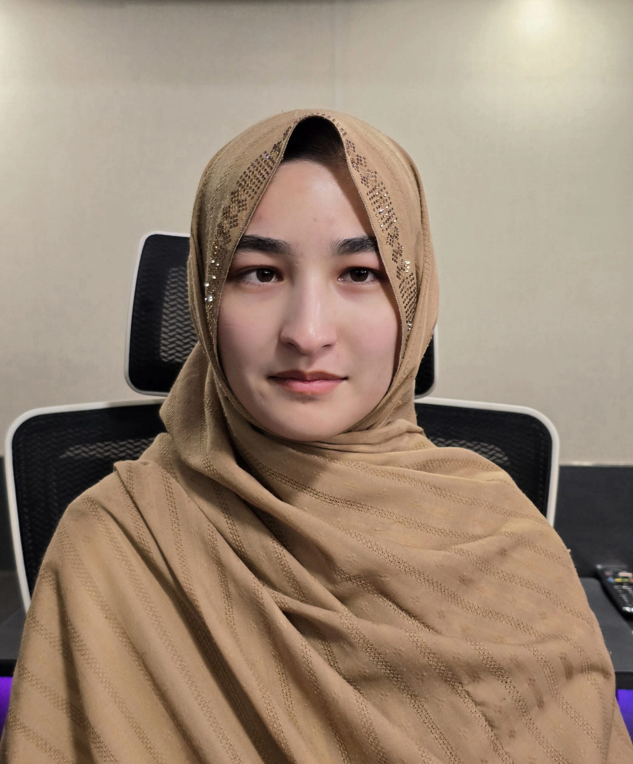 A woman wearing a tan hijab sits in a black office chair, posing for a photo that showcases her website design skills