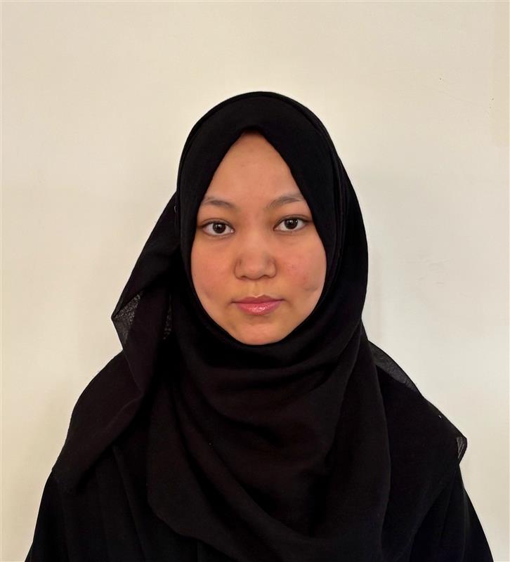 A woman in a black hijab with a neutral expression, wearing a black outfit, against a white background, with a focus on