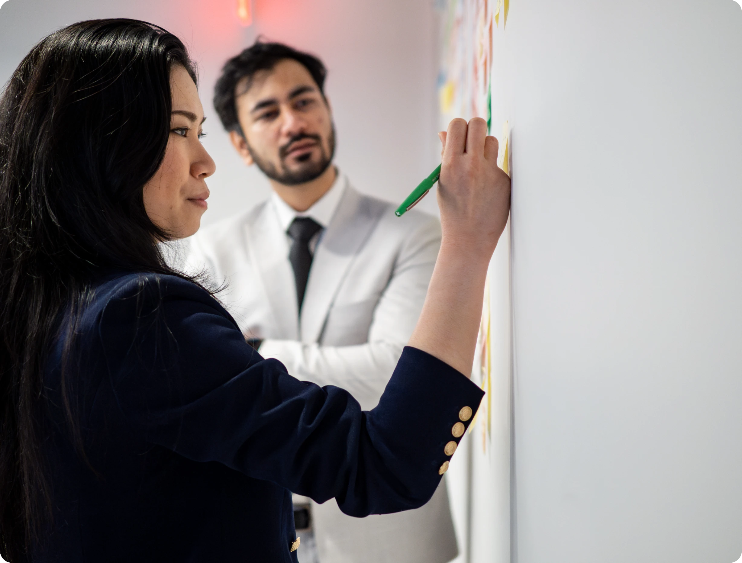 A woman with long black hair and a man with a beard and white suit writing on a whiteboard with a green marker, both in a